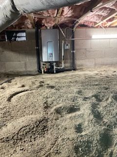 A water heater installed in a crawl space with exposed insulation, cinder block walls, and a dirt floor.