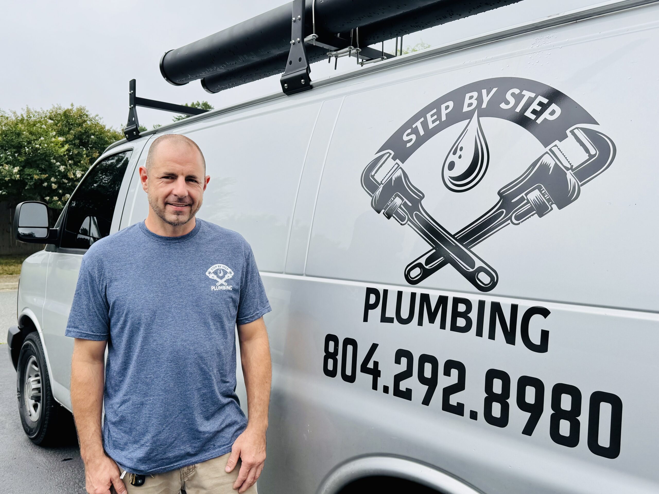 A man in a blue t-shirt stands next to a white van with Step By Step Plumbing and a phone number printed on the side.