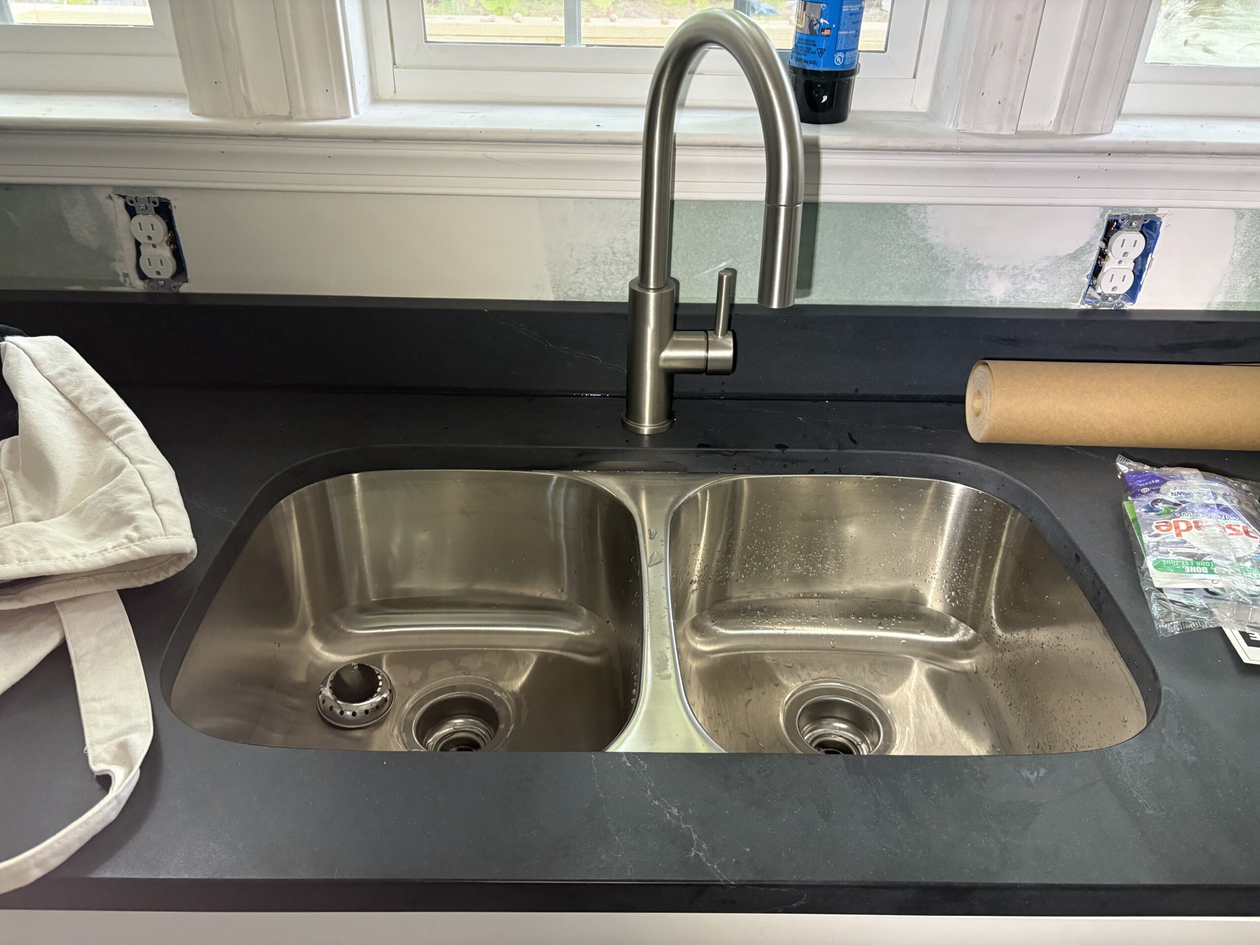 A double-basin stainless steel kitchen sink with a modern faucet, set in a dark countertop—perfect for fixture repair and replacement. Nearby are a white cloth bag, a paper towel roll, and some packaged items.