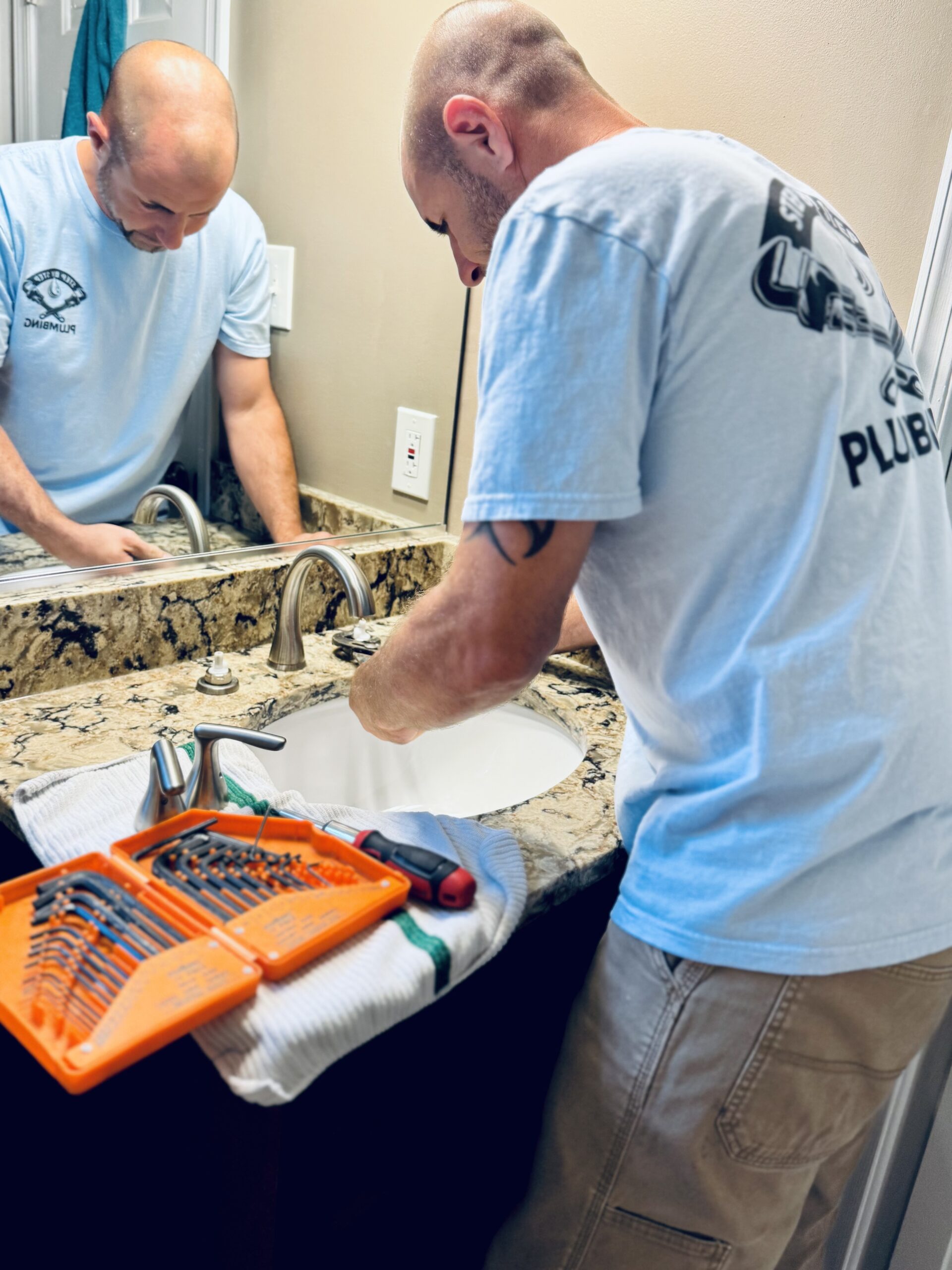 A man repairs a bathroom sink, using tools laid out on a towel over the granite countertop.