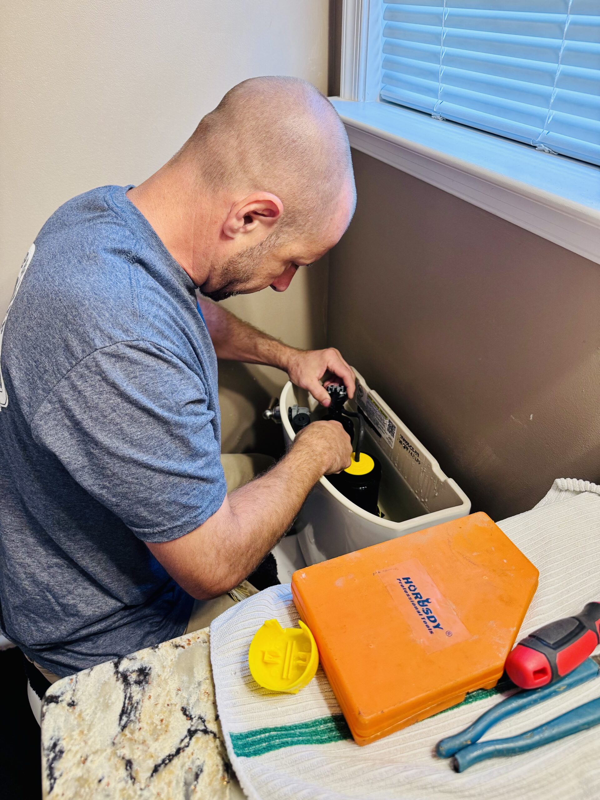 A person is repairing a toilet tank, handling the internal components. Tools and parts are placed on a nearby countertop.
