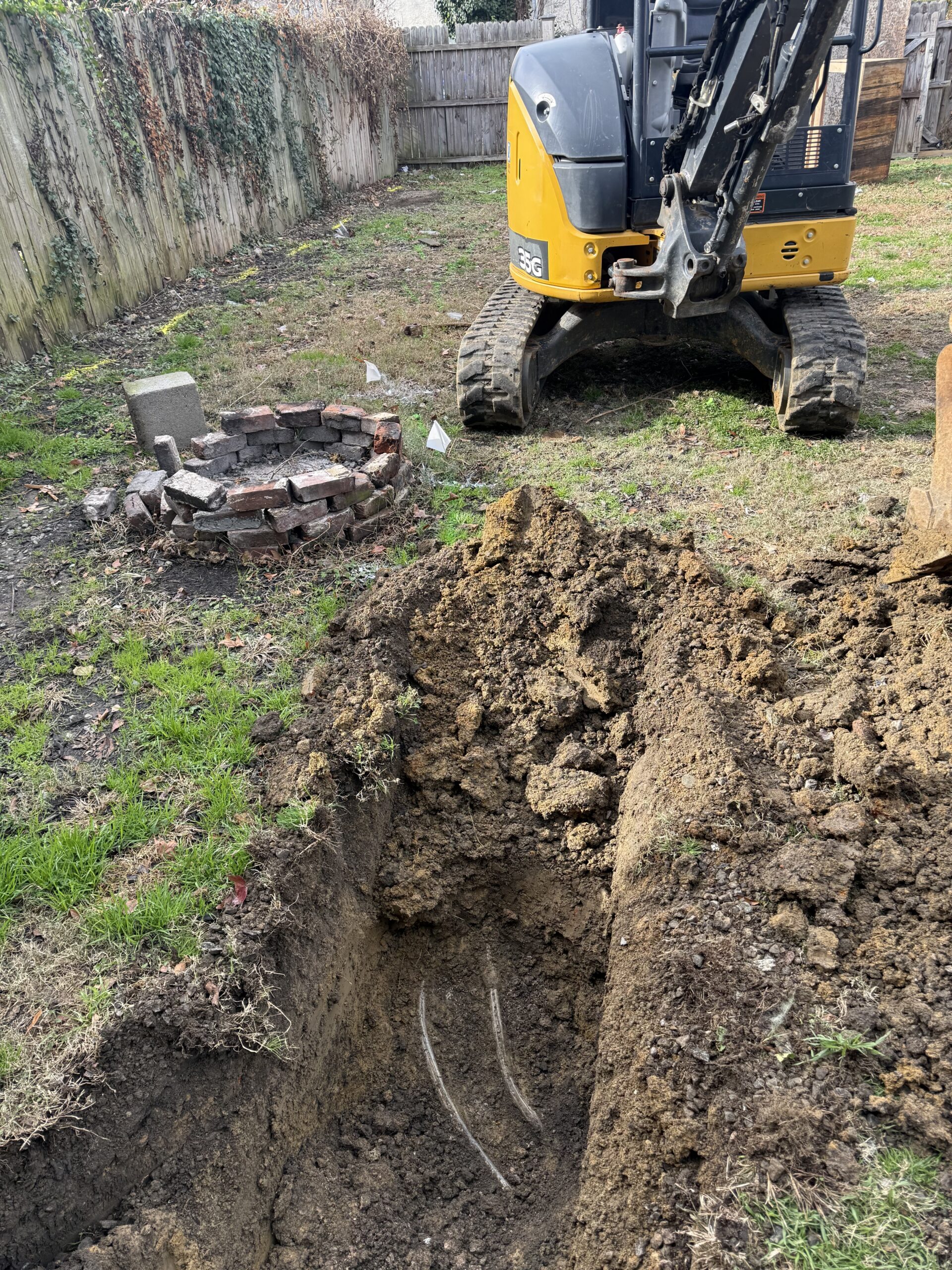A small excavator digs a trench in a backyard for sewer inspection, repair, and replacement, revealing exposed cables in the soil; a circular brick structure and utility markers are visible nearby.