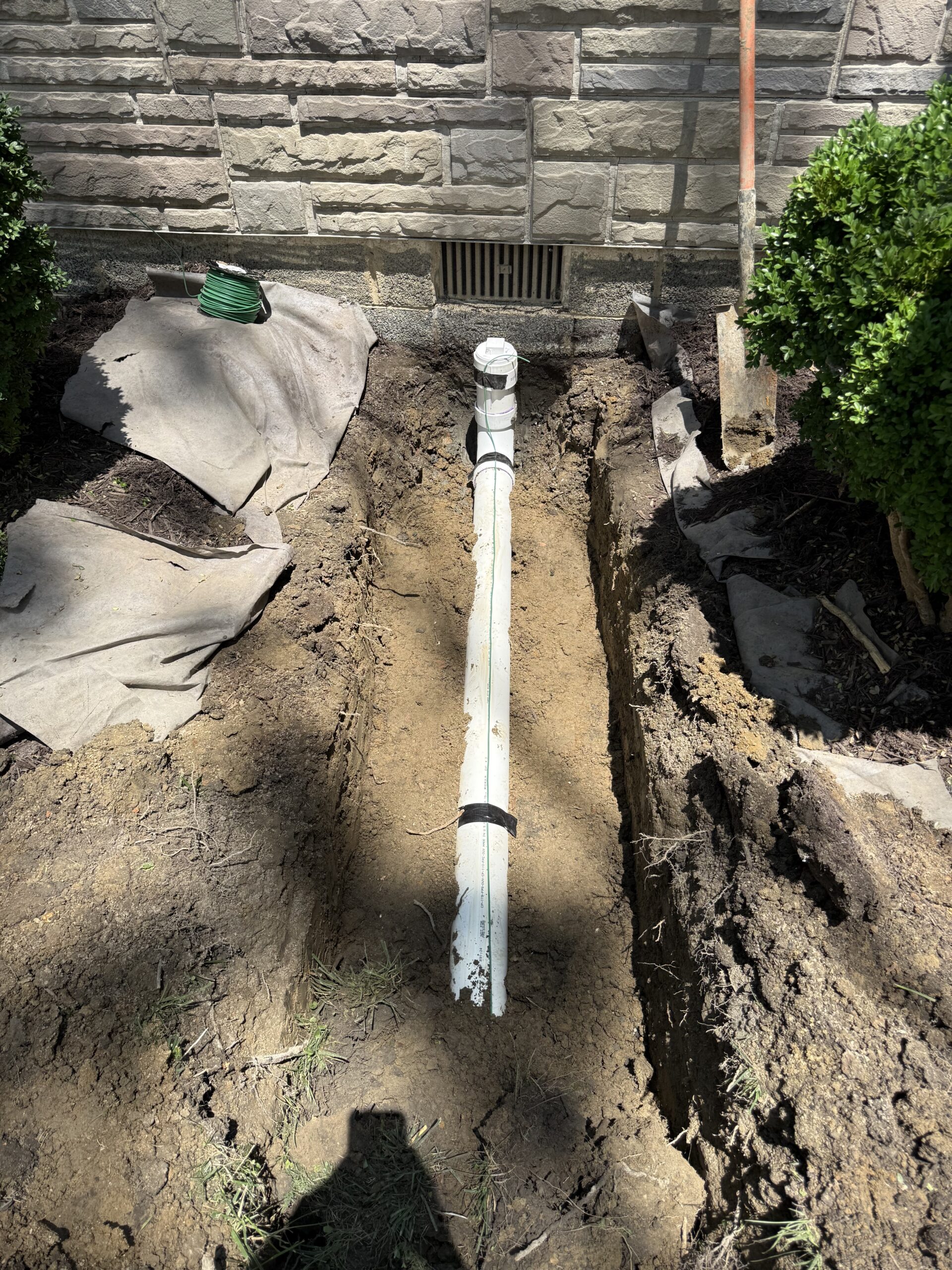 A white drainage pipe is partially buried in a trench alongside a stone building, with a shovel, hose, and landscaping fabric visible nearby.