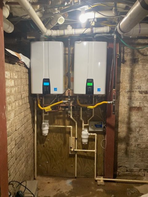 Two wall-mounted tankless water heaters with digital displays and connected piping are installed in a basement with exposed brick walls and overhead plumbing.